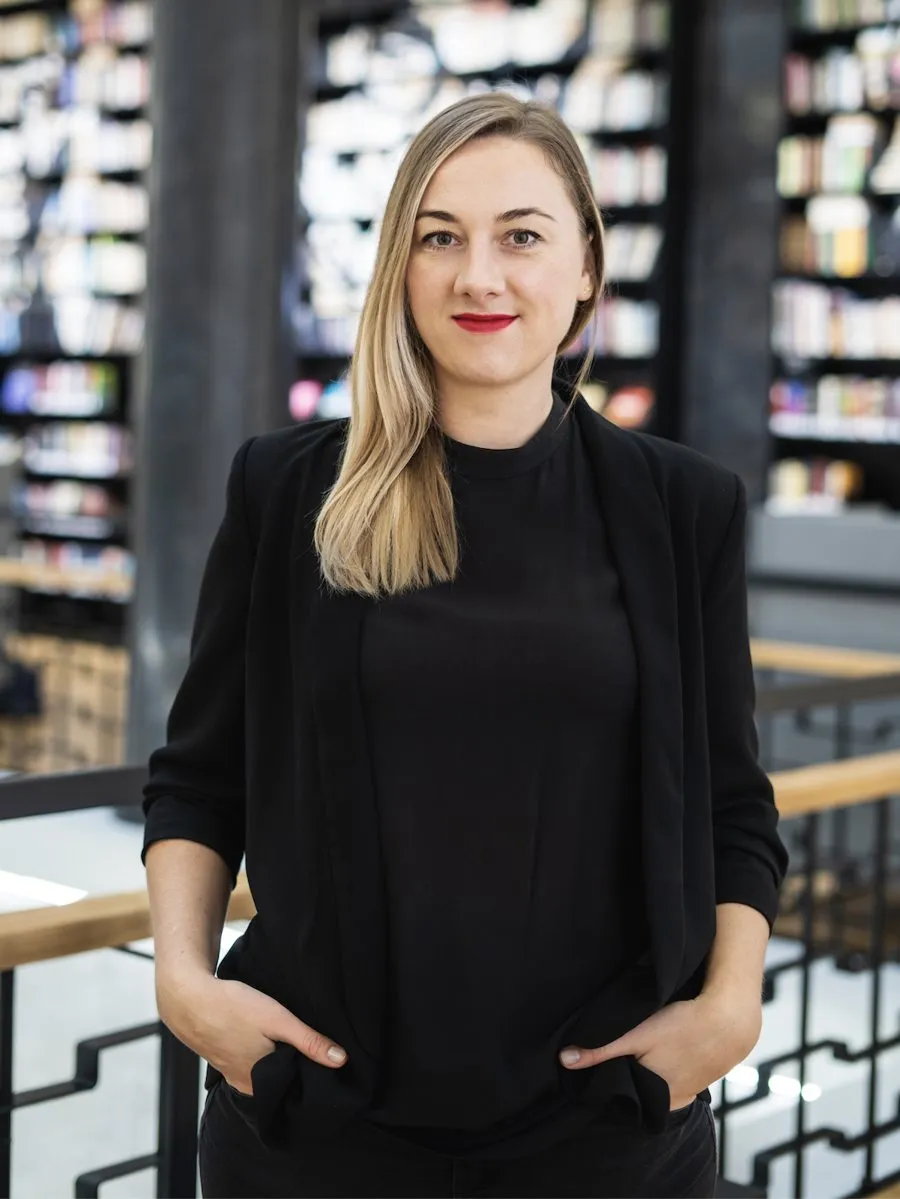 Dr. Dalia Bagdžiūnaitė, juodu švarku, stovi bibliotekoje prie knygų lentynų